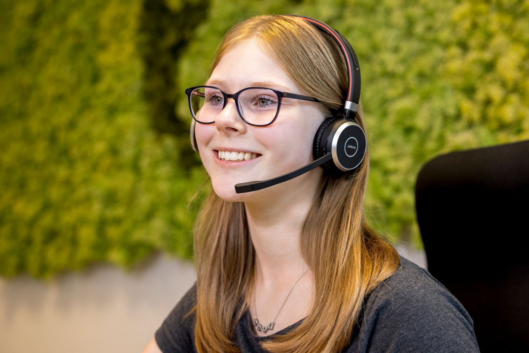 Eine junge Frau mit langen blonden Haaren und Brille, die ein Headset mit Mikrofon trägt, sitzt lächelnd in einem Bürostuhl. Im Hintergrund ist eine grüne, mit Pflanzen bewachsene Wand zu sehen.